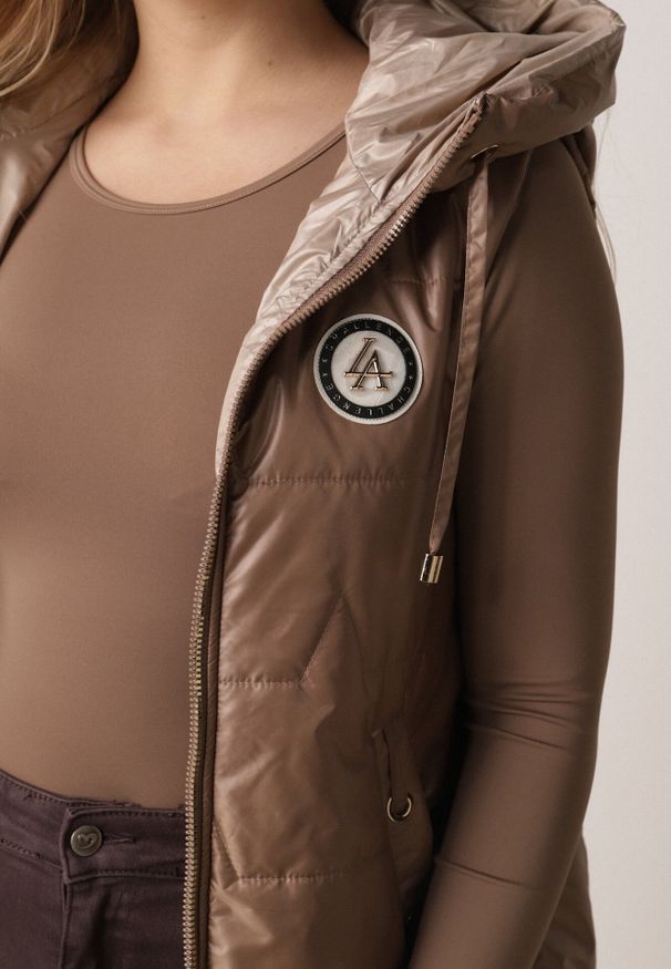 Born2be - Jasnobrązowa Pikowana Kamizelka o Długim Kroju z Nieodpinanym Kapturem i Aplikacją Koretes. Okazja: na co dzień. Kolor: brązowy. Materiał: prążkowany, poliester. Długość: długie. Wzór: aplikacja. Styl: casual