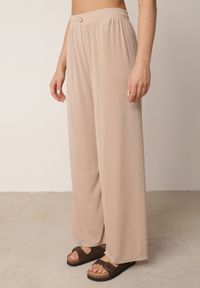 Born2be - Beżowe Spodnie z Wysokim Stanem i Szerokimi Nogawkami Zdobione Guzikiem Kelesh. Okazja: do pracy, na co dzień. Stan: podwyższony. Kolekcja: plus size. Kolor: beżowy. Materiał: tkanina, prążkowany. Długość: długie. Wzór: aplikacja. Styl: elegancki, casual #1