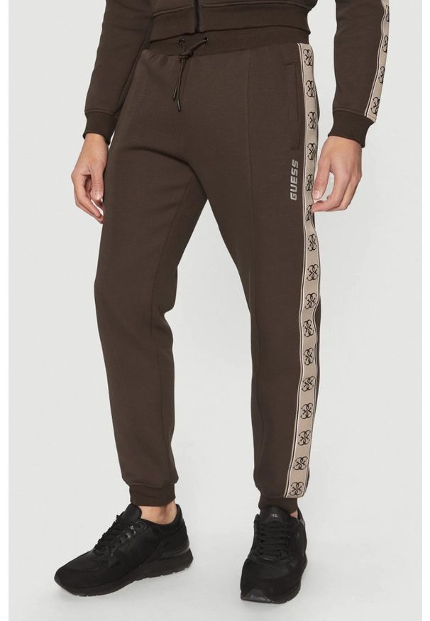 Guess - GUESS Brązowe męskie spodnie dresowe z lampasami Mickey, Rozmiar S. Kolor: brązowy. Materiał: dresówka