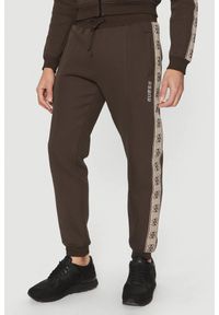 Guess - GUESS Brązowe męskie spodnie dresowe z lampasami Mickey, Rozmiar S. Kolor: brązowy. Materiał: dresówka #1